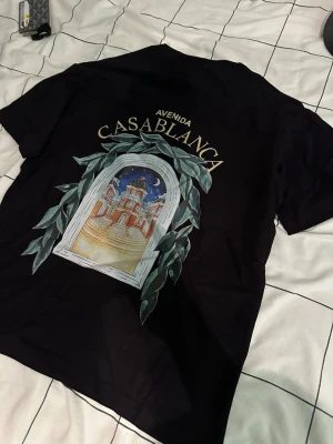 Svart Casablanca Avenida t-shirt - Snygg svart t-shirt från Casablanca med trycket 'Avenida Casablanca' och ett färgglatt motiv av en byggnad och nattmotiv omgiven av gröna blad både fram och bak. Klassisk passform och rund hals. Perfekt för dig som vill sticka ut med stil.