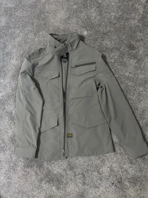 Grå utilityjacka från G-Star RAW - Snygg grå utilityjacka från G-Star RAW med hög krage, dragkedja framtill och flera praktiska fickor med lock och dragkedja. Tillverkad i ett slitstarkt material som passar perfekt till streetwear och vardagsstil.
