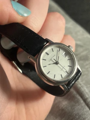 Certina silverklocka med svart läderband - Stilren Certina armbandsklocka med rund silverfärgad boett, vit urtavla och datumvisning. Svart läderarmband och minimalistisk design med tydliga markeringar. Perfekt för dig som gillar klassiska accessoarer.