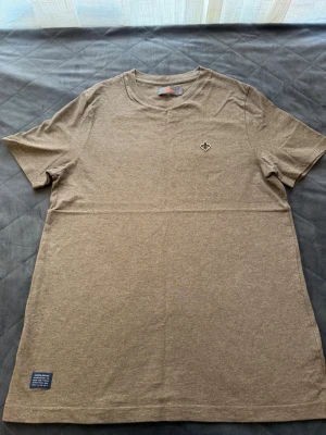 Beige t-shirt från Morris - Säljer en beige t-shirt från Morris Stockholm med liten logga på bröstet och patch vid nederkanten. Klassisk rund hals och korta ärmar. Tillverkad i mjuk bomull för skön känsla och enkel stil.