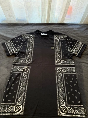 Svart bandanamönstrad t-shirt Plein - Snygg svart t-shirt från Plein med unikt bandanamönster i vitt. T-shirten har korta ärmar, rund halsringning och ett diskret Plein-emblem på bröstet. Tillverkad i mjuk bomull som känns skön mot huden. Perfekt för dig som vill sticka ut med en cool streetwear-look.