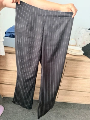 Svarta kostymbyxor med vita ränder - Säljer ett par svarta kostymbyxor med tunna vita kritstrecksränder. Byxorna har rak passform och hög midja, vilket ger en clean och klassisk look. Materialet är en mjuk och lite glansig mix, perfekt för en stilren outfit.