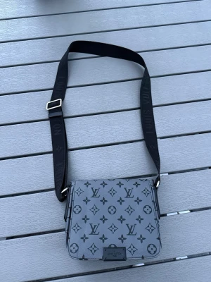 Grå axelväska  - Snygg grå axelväska  med klassiskt monogrammönster i svart. Väskan har justerbar svart axelrem med logga och silverfärgade detaljer. Tillverkad i tåligt konstläder och har en stilren, fyrkantig form som ger en modern vibe.