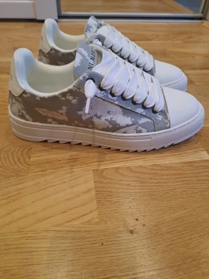 Vita och grå sneakers Numeris - Snygga sneakers från Numeris i vitt och grått camomönster. Skorna har tjocka vita snören, vit tå och sula samt en låg profil. Perfekta för dig som gillar streetwear och vill sticka ut lite extra.