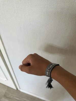 Dior armband - Svart tygarmband från Christian Dior med vit broderad text 'Christian Dior J'Adior'. Armbandet har justerbar knytning och tofsar i ändarna för en trendig och personlig stil. Perfekt accessoar för dig som gillar designerplagg och vill sticka ut.