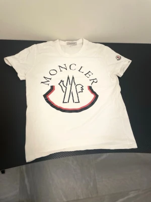 Vit Moncler t-shirt med stor logga - Snygg vit t-shirt från Moncler med stor logga och grafiskt tryck på bröstet. Klassisk rund halsringning och korta ärmar. Ikonisk Moncler-patch på ena ärmen. Tillverkad i mjuk bomull för skön känsla.