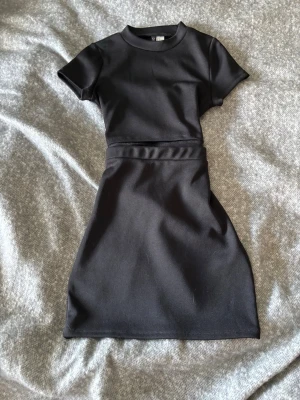 Svart kortärmad tvådelad klänning - En stilren svart klänning med korta ärmar och rund hals. Klänningen har en figurnära passform upptill och en lätt utsvängd kjol. Perfekt för dig som gillar enkel och modern stil.