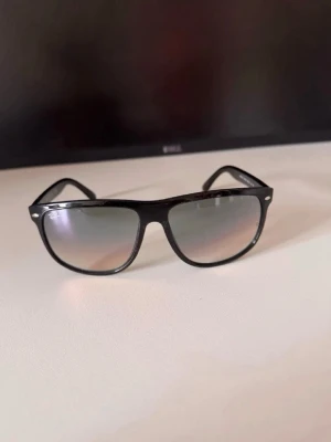 Svarta Ray-Ban solglasögon - Snygga svarta solglasögon från Ray-Ban med klassisk boyfriend-form. Bågarna är blanka och har Ray-Ban-loggan på sidan. Glasen är mörka och ger en cool look till alla outfits.