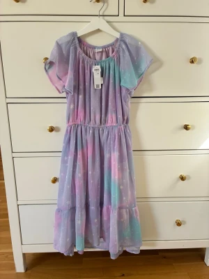 Klänning barn Lindex  - Söt långklänning i pastellfärger med lila, rosa, blå och turkosa toner. Klänningen har ett stjärnmönster, korta ärmar och resår i midjan för en bekväm passform. Oanvänd med prislapp kvar.  