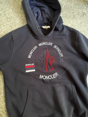 Mörkblå Moncler hoodie med brodyr - Snygg mörkblå hoodie från Moncler med stor broderad logga och text på bröstet i vitt, rött och blått. Klassisk känguruficka framtill och Moncler-märke på ärmen. Tillverkad i mjukt bomullsmaterial och har en stor huva för extra komfort.