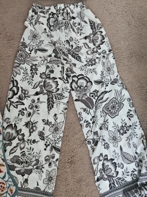 Set med blommigt mönster i svartvitt - Säljer ett snyggt set med vida byxor och matchande off-shoulder topp. Setet är vitt med svart blommigt mönster och har elastisk midja och topp för skön passform. Perfekt för dig som gillar bohemisk stil och vill sticka ut med ett unikt mönster.