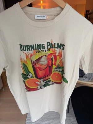 Vit Burning Palms t-shirt Vailent - Säljer en vit t-shirt från Vailent med färgglad Burning Palms Beach Bar-print framtill. Motivet visar en drink med is, apelsiner och eldiga detaljer. Klassisk rund hals och korta ärmar. Perfekt för dig som gillar statement-plagg och coola tryck.