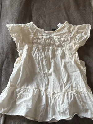 Vit blus med volang och kort ärm - Söt vit blus med volangkant nedtill och korta ärmar. Blusen har dekorativa sömmar och lätt rynkade detaljer upptill. Tillverkad i ett tunt, luftigt tyg som passar perfekt till sommaren. Enkel att matcha med både jeans och kjol.