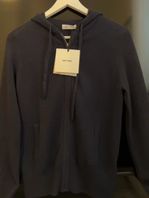 Mörkblå hoodie från Soft Goat - Mörkblå hoodie från Soft Goat i mjuk kashmir. Dragkedja framtill, två fickor och justerbar huva med snören. Perfekt för dig som vill ha en stilren och bekväm hoodie i premiumkvalitet. Enkel design utan tryck eller mönster.