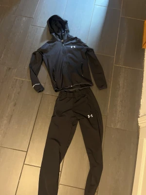 Svart träningsset från Under Armour - Snyggt svart träningsset från Under Armour med huva och dragkedja på jackan samt matchande byxor. Setet har vita loggor på både jacka och byxa, samt praktiska fickor med dragkedja. Perfekt för gymmet eller löprundan.