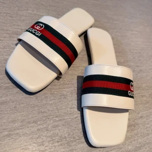 Vita slip-in skor tofflor el sandaler stl 39 med snyggt, trendigtband - Säljer ett par vita sandaler, slip-in skor el tofflor med breda band i klassiska gröna och röda ränder samt logga. Stl 39 De är lika fina till sommarklänningen som jeansshortsen. Sandalerna har fyrkantig tå och platt sula. Perfekta för dig som vill ha en clean och lyxig look till sommaren.