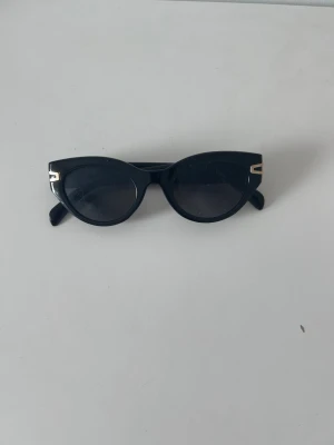 Svarta cat eye solglasögon - Snygga svarta solglasögon i klassisk cat eye-modell med mörka glas och guldfärgade detaljer vid skalmarna. Perfekta för att lyfta vilken outfit som helst och ger en cool retrovibe. Ramen är i plast och glasen är mörkt tonade.