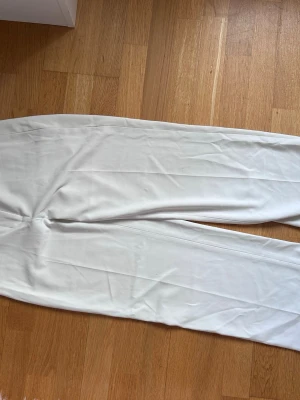 Vita raka kostymbyxor - Säljer ett par stilrena vita kostymbyxor med rak passform och pressveck längs benen. Byxorna har en klassisk look och är perfekta för en clean och modern outfit. Materialet ser ut att vara mjukt och följsamt.