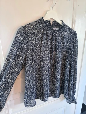Blommig blus från Holly & Whyte - Mönstrad blus från Holly & Whyte i marinblått med vita blommor. Blusen har långärmad design, hög krage med volang och mjukt, tunt tyg som ger en luftig känsla. Perfekt för dig som gillar romantiska detaljer och stilren look.
