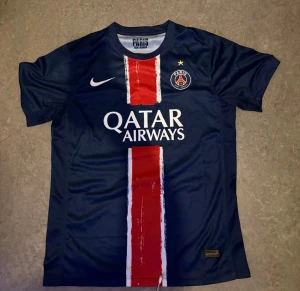 PSG matchtröja blå Nike - Snygg blå PSG matchtröja från Nike med röd och vit rand i mitten, klubbmärke och Qatar Airways-tryck på bröstet. Tillverkad i lätt och ventilerande material, perfekt för fotboll eller träning. Rund hals och korta ärmar.