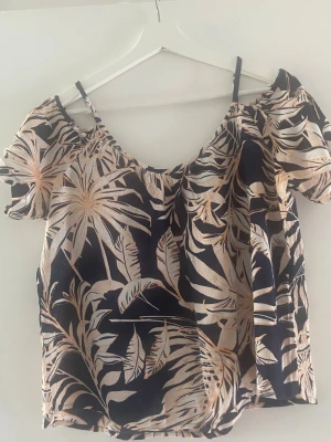 Blommig offshoulder topp i marinblått - Snygg offshoulder topp med marinblå botten och stort blommönster i vit och ljusorange. Toppen har tunna axelband och korta puffärmar. Perfekt för sommarens alla tillfällen och ger en fräsch vibe till din outfit.