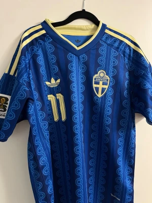  Sverige matchtröja Elanga 11 - Säljer en blå Sverige matchtröja från Adidas med Elanga och nummer 11 på ryggen. Tröjan har gula detaljer, randigt mönster och Sveriges landslagsemblem på bröstet. Klassiska Adidas-ränder på axlarna och FIFA-märke på ärmen. Perfekt för fotbollsfans!