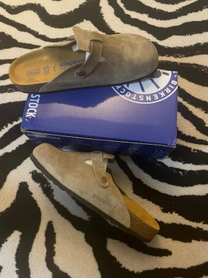 Birkenstock Boston mockatofflor beige - Säljer ett par Birkenstock Boston tofflor i beige mocka med justerbart spänne och korksula. Klassisk slip-in modell som är både stilren och bekväm. Perfekta för dig som gillar en avslappnad look och vill ha något tidlöst till garderoben.