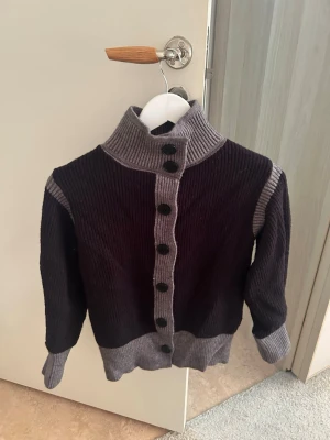 Svart tjock kofta med grå detaljer - Snygg svart kofta med grå ribbstickade detaljer längs knäppningen, kragen, ärmsluten och nederkanten. Koftan har hög krage och stora svarta knappar framtill ♠️🩶