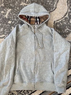 Grå zip hoodie från Burberry - Snygg grå zip hoodie från Burberry med klassiskt rutigt foder i huvan och dragsko. Hoodien har två fickor framtill och ribbade muddar vid ärmar och nederkant. Perfekt för en avslappnad och stilren look.