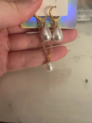 Guldörhängen med droppformade pärlor - Säljer ett par snygga örhängen med guldfärgade ringar och droppformade vita pärlor. Ena örhänget har en extra kedja med en tredje pärla för en cool asymmetrisk look. Perfekta för dig som gillar trendiga accessoarer med lite edge.