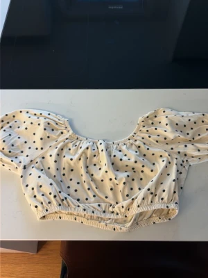 Prickig croppad offshoulder topp - Söt croppad topp i vitt med svarta prickar. Modellen är offshoulder med puffiga korta ärmar och resår vid halsringning och midja. Perfekt för dig som gillar trendiga och lekfulla plagg.Storlek 152cm. 11-12 år 