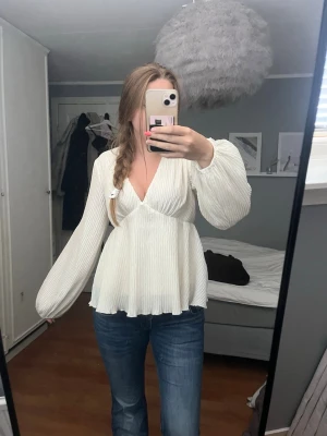 Vit plisserad blus med v-ringning - Säljer en superfin vit blus med plisserat tyg och v-ringning. Blusen har långa, vida ärmar och en lätt utsvängd passform som ger en flowy känsla. Perfekt att styla med jeans eller kjol för en trendig look.