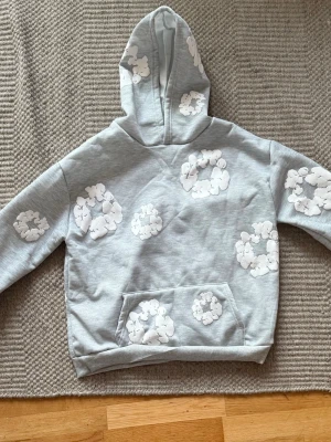 Ljusgrå hoodie från denim tears  - Säljer en ljusgrå hoodie med huva från denim tears, tryck över hela tröjan. Hoodien har en klassisk känguruficka framtill och ribbade muddar vid ärmslut och nederkant. Perfekt för dig som gillar en unik och avslappnad 