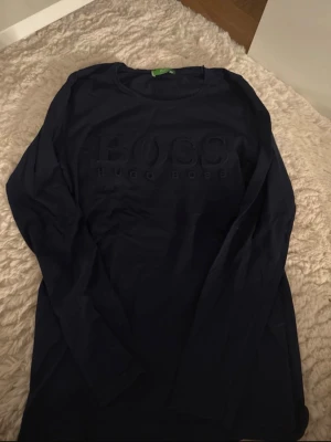 Svart långärmad tröja Hugo Boss - Snygg svart långärmad tröja från Hugo Boss med diskret broderad logga på bröstet. Tröjan har rund halsringning och är tillverkad i mjukt material som känns skönt mot huden. Perfekt för en stilren och avslappnad look.