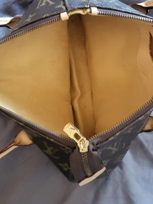 Louis Vuitton monogram handväska - Snygg handväska från Louis Vuitton i klassiskt brunt monogrammönster med beige insida. Väskan har dragkedja i metall och handtag i ljust läder. Perfekt för dig som gillar lyxiga accessoarer och tidlös design.