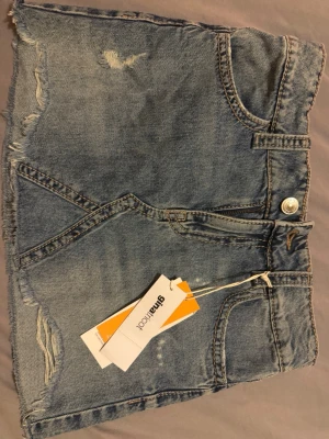 Blå jeanskjol med rå kant - Trendig blå jeanskjol med rå, fransig kant och slitningar framtill. Kjolen har klassiska fickor både fram och bak med snygga detaljer och knappar. Perfekt för en avslappnad och cool stil. 
