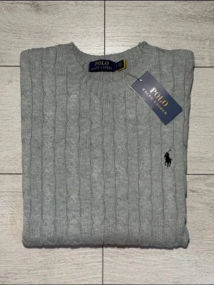 Grå kabelstickad tröja Polo Ralph Lauren - Snygg grå kabelstickad tröja från Polo Ralph Lauren med klassisk rund halsringning och broderad logga på bröstet. Tröjan är i storlek S (lutar mer åt Xs) och passar personer som är mellan 165-175. Skick 10/10 då den är helt oanvänd! Nypris: 2600kr
