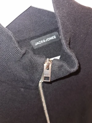 Svart ziptröja från Jack & Jones - Svart stickad ziptröja från Jack & Jones med hög krage och hel dragkedja framtill. Tröjan har långa ärmar och två fickor framtill. Perfekt för en stilren och avslappnad look.