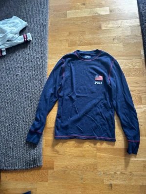 Mörkblå pyjamastopp Polo Ralph Lauren - Mörkblå långärmad pyjamastopp från Polo Ralph Lauren med röd kontrastsöm och liten amerikansk flagga samt Polo-tryck på bröstet. Tillverkad i mjukt, strukturerat material som är perfekt för mysiga kvällar.
