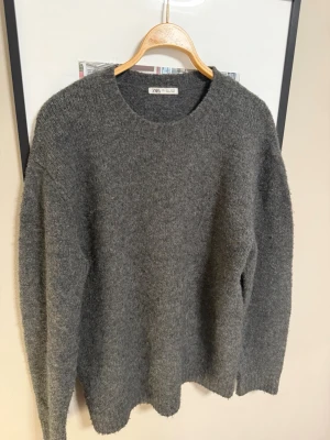 Mörkgrå stickad tröja från Zara - Mysig mörkgrå stickad tröja från Zara med rund halsringning och lång ärm. Tröjan har en enkel, klassisk design och är tillverkad i ett mjukt, fluffigt material som passar perfekt för kyligare dagar.