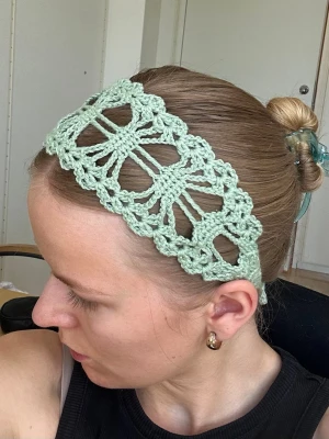 Handvirkat ljusgrönt hårband - Säljer ett handvirkat hårband i ljusgrönt garn. Hårbandet har ett brett, luftigt mönster och knyts med långa snören i nacken. Perfekt för dig som vill ha en unik accessoar med boho-vibe och cottage core känsla.