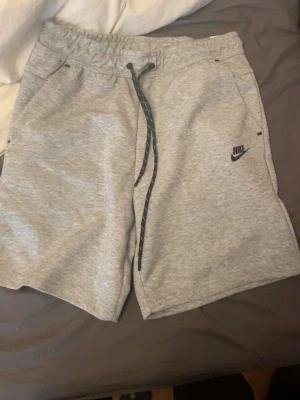 Gråa Nike tech shorts - Säljer ett par gråa Nike shorts med svart snörning i midjan och diskret logga på vänster ben. Shortsen har två sidofickor och en bakficka med dragkedja. Perfekta för chill dagar på stranden eller träning på gymmet, även om du vill bara sitta hemma och tar de lugnt 🌟