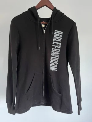 Harley-Davidson damhoodie med dragkedja - Harley-Davidson damhoodie med dragkedja Storlek: M Passform: Slim Fit Krage: Huva med dragsko Material: Bomull & Polyester  Mått: Bröstvidd 100 cm, längd 70 cm, axel 48 cm Skick: Denna hoodie är i ett väldigt fint skick utan defekter  Nypris cirka: 700 kr