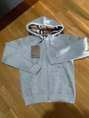 Grå zip hoodie från Burberry - Snygg grå zip hoodie från Burberry med klassiskt rutigt foder i huvan. Tillverkad i mjukt material med dragsko i huvan och två fickor framtill. Perfekt för en avslappnad och trendig look. Komm med bud vid snabb affär 