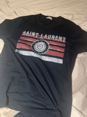 Svart Saint Laurent t-shirt - Snygg svart t-shirt från Saint Laurent med tryck i vitt och rött framtill. T-shirten har klassisk rund hals och korta ärmar. Materialet är mjuk bomull som känns skönt mot huden. Perfekt för dig som vill ha en stilren och trendig look.
