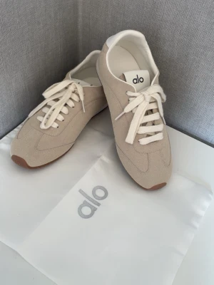 ALO skor i mocka  - Snygga beige sneakers från Alo med ovandel i mocka och vita detaljer. Skorna har vita snören, vit insida och en klassisk låg siluett med gummisula i brunt. Perfekta för dig som gillar stilrena och trendiga sneakers.