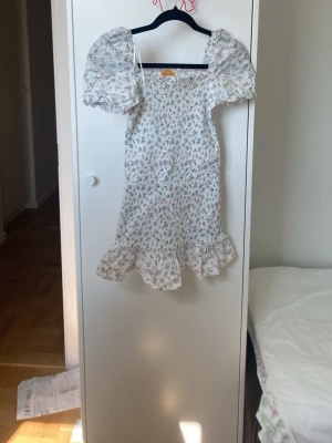 Vit blommig kortklänning från Gina trikot - Finns INTE i butik längre! Strechig och supersöt vit kortklänning från Gina tricot med små blommor i rosa, blått och grönt. Klänningen har puffärmar, volangkant nertill och smockad överdel som ger en bekväm passform. 