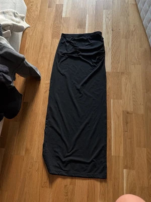 Svart långkjol med slits och rynk - Säljer en svart långkjol med snygg rynkdetalj i midjan och en slits framtill. Kjolen har en rak passform och är gjord i ett mjukt, stretchigt material som känns bekvämt. Perfekt att styla med en crop top eller oversized tröja.