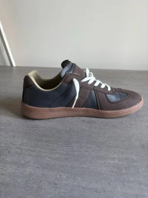 Bruna och grå sneakers med vit sula - Snygga sneakers i brunt och grått med vita skosnören och beige insida. Skon har en klassisk låg siluett och ovandelen är i en mix av mocka och slätt material. Yttersulan är ljusbrun och ger en retrovibe. Perfekt för dig som gillar stilrena och tidlösa sneakers.
