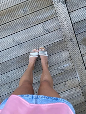 Vita sandaler med ränder - Säljer ett par vita sandaler med två breda remmar över foten och diskreta ränder. Sandalerna har öppen tå och platt sula, perfekta för varma dagar. Passar till både jeansshorts och klänning.